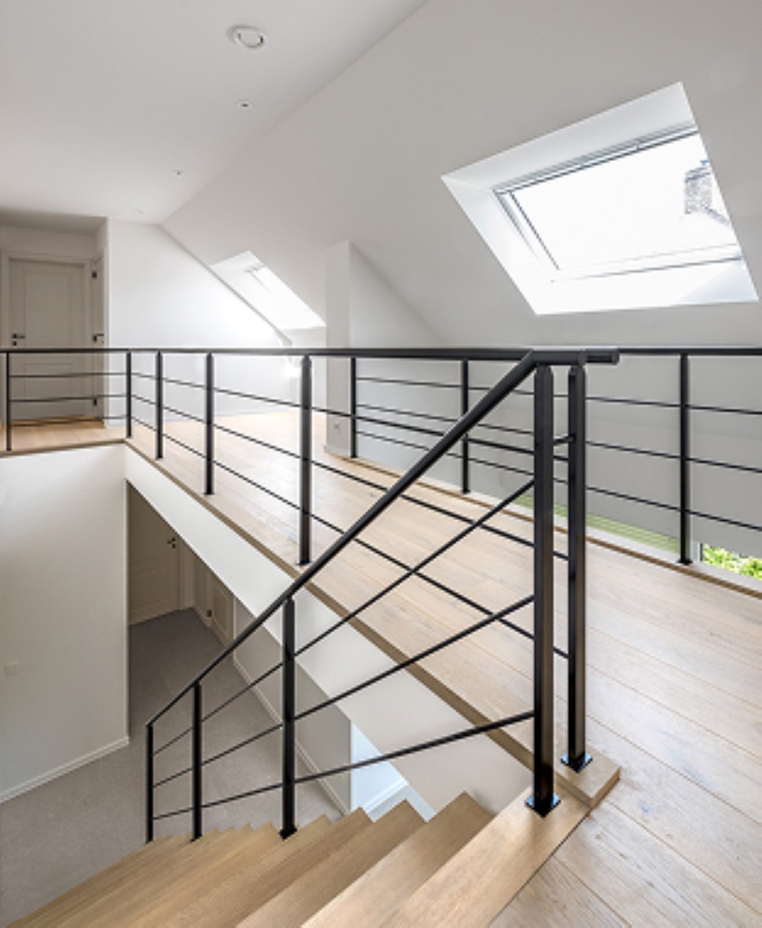 Lichtrijke bovenverdieping met houten vloer, zwarte metalen trapleuning en schuine plafonds met grote dakramen die natuurlijk licht binnenlaten.